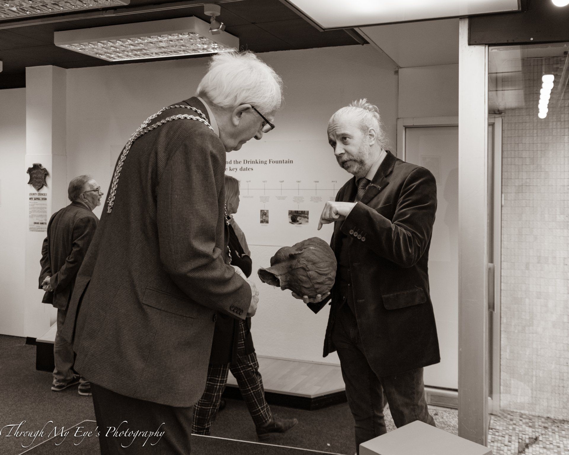 Man in jacket shows object to man in formal attire. Inside a building, other people nearby.