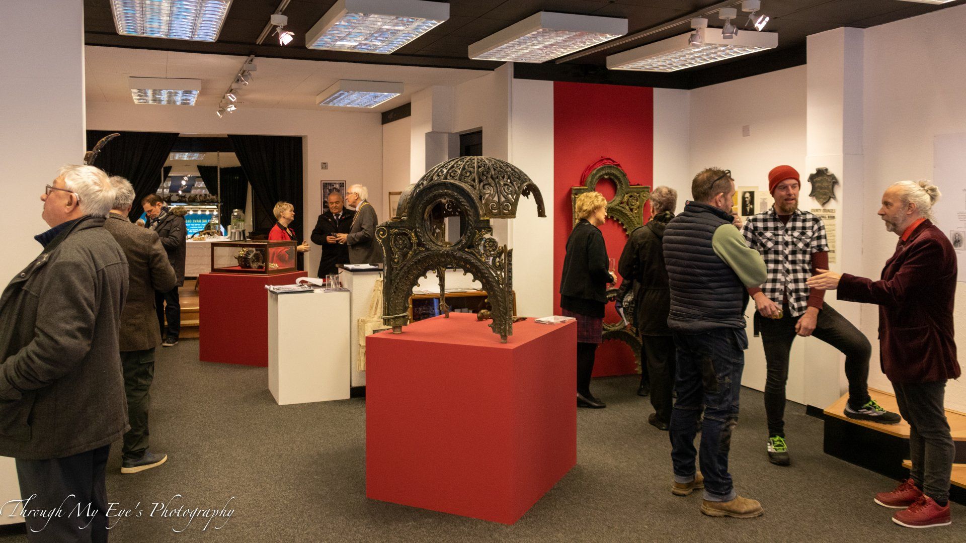 People in an art gallery viewing sculptures and paintings on display; red and white walls, gray carpet.