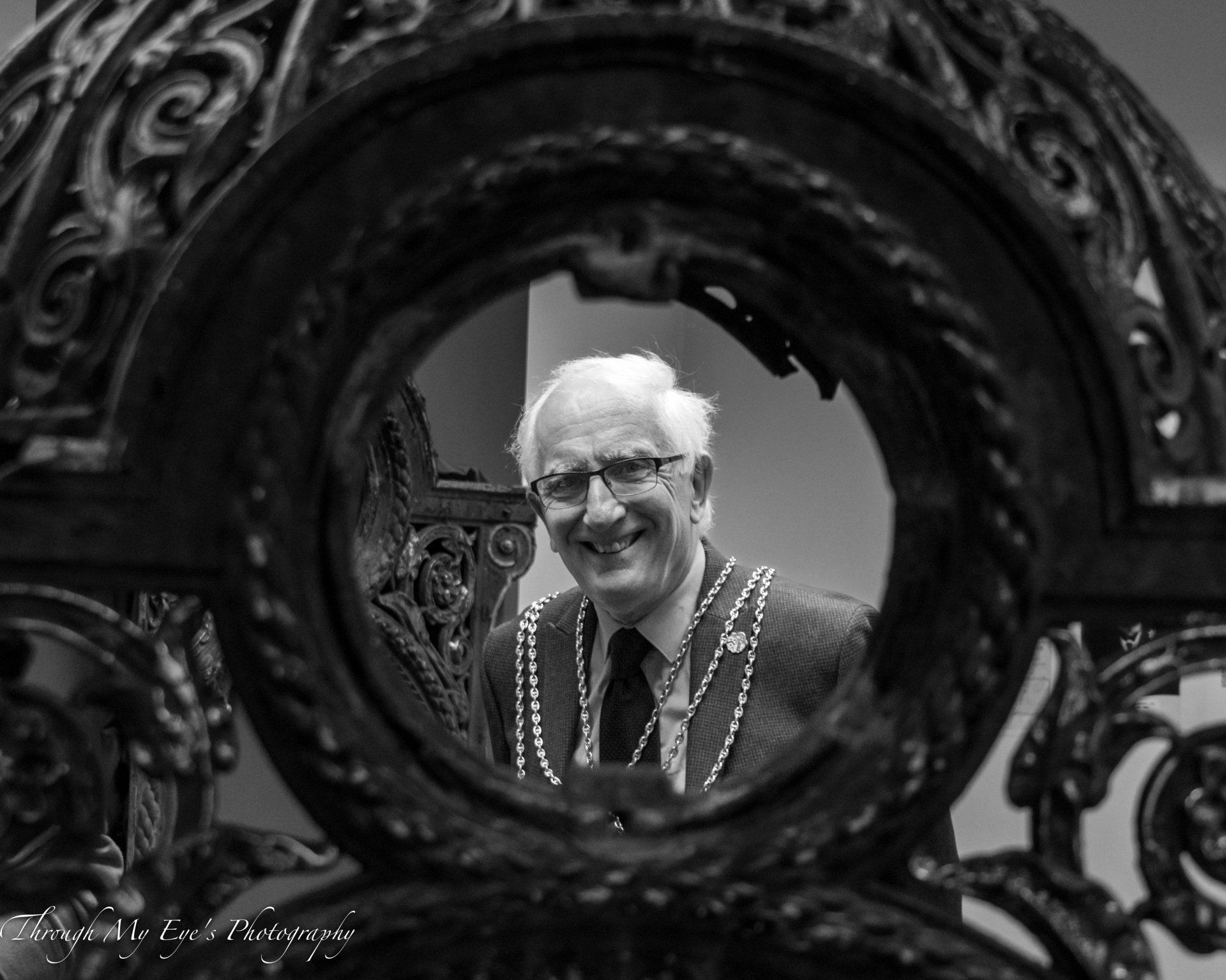 Man with glasses and chain, smiling, viewed through ornate ironwork.
