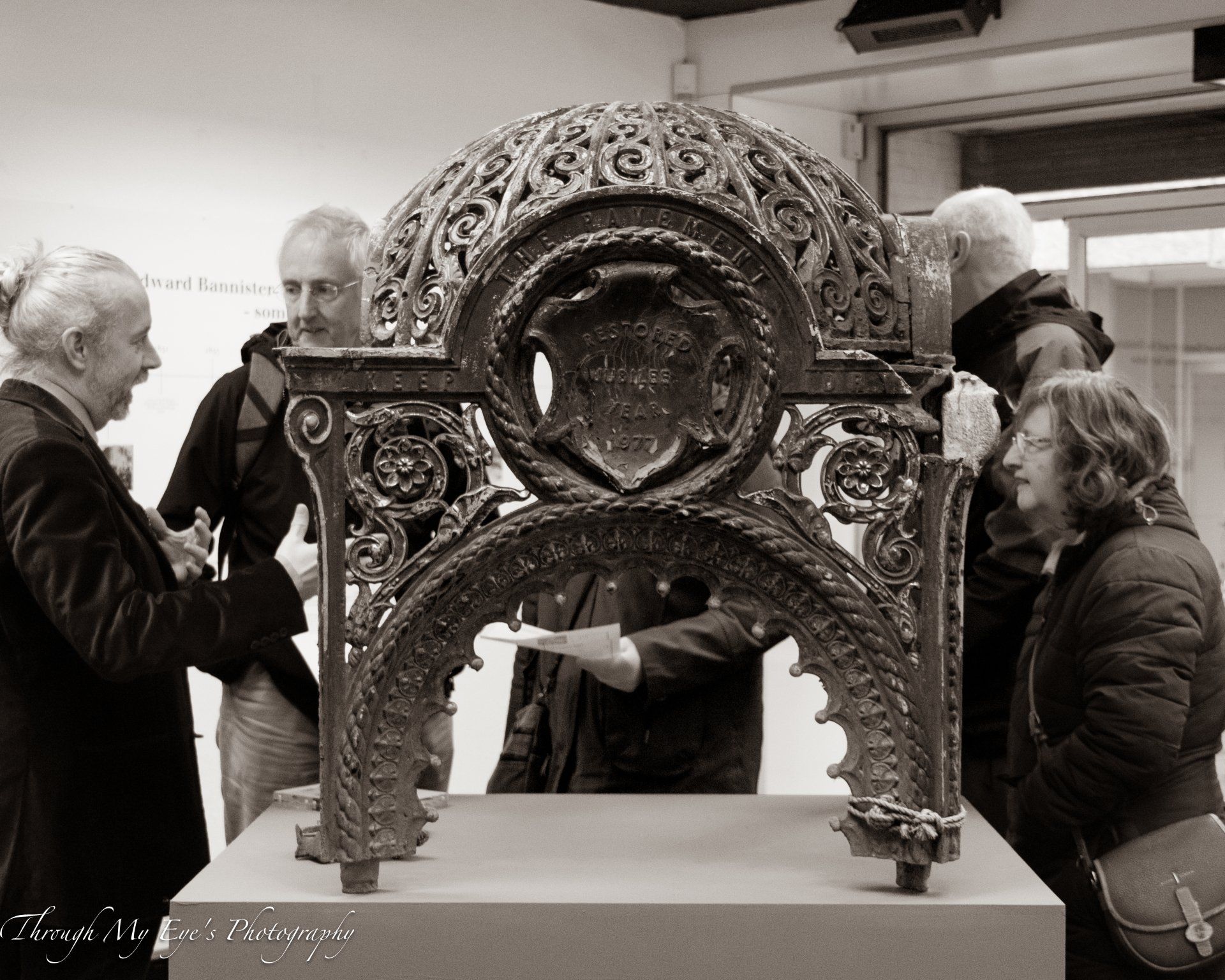 People examine an ornate, arched metal structure with a crest on display in a gallery.