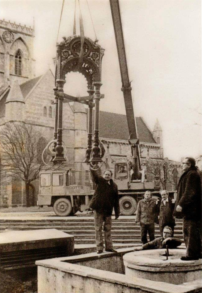 Crane lifts ornate fountain structure; men watch near building in town square.