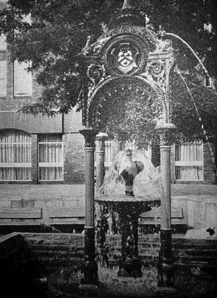 Ornate fountain with water cascading, in front of a brick building.