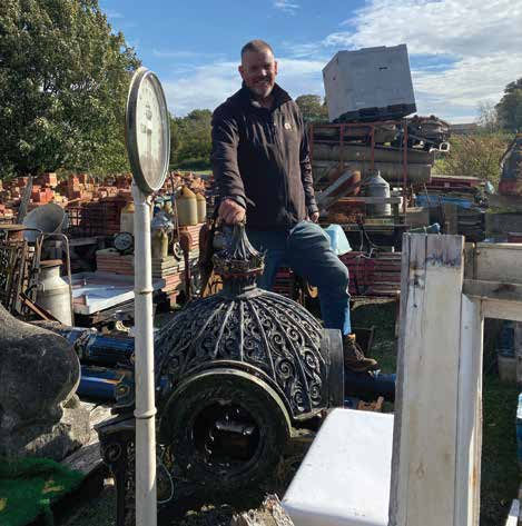 Man in brown jacket, standing among salvage yard objects, holds ornate metal object, scale visible.