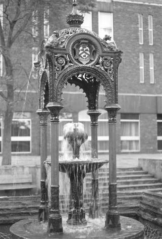 Ornate fountain with water cascading, in front of a brick building.