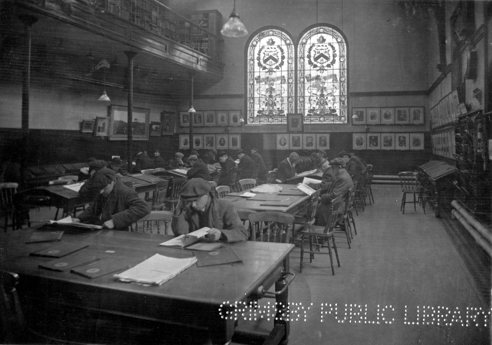 Men seated at desks, reading in a library. Stained glass windows and artwork on walls.