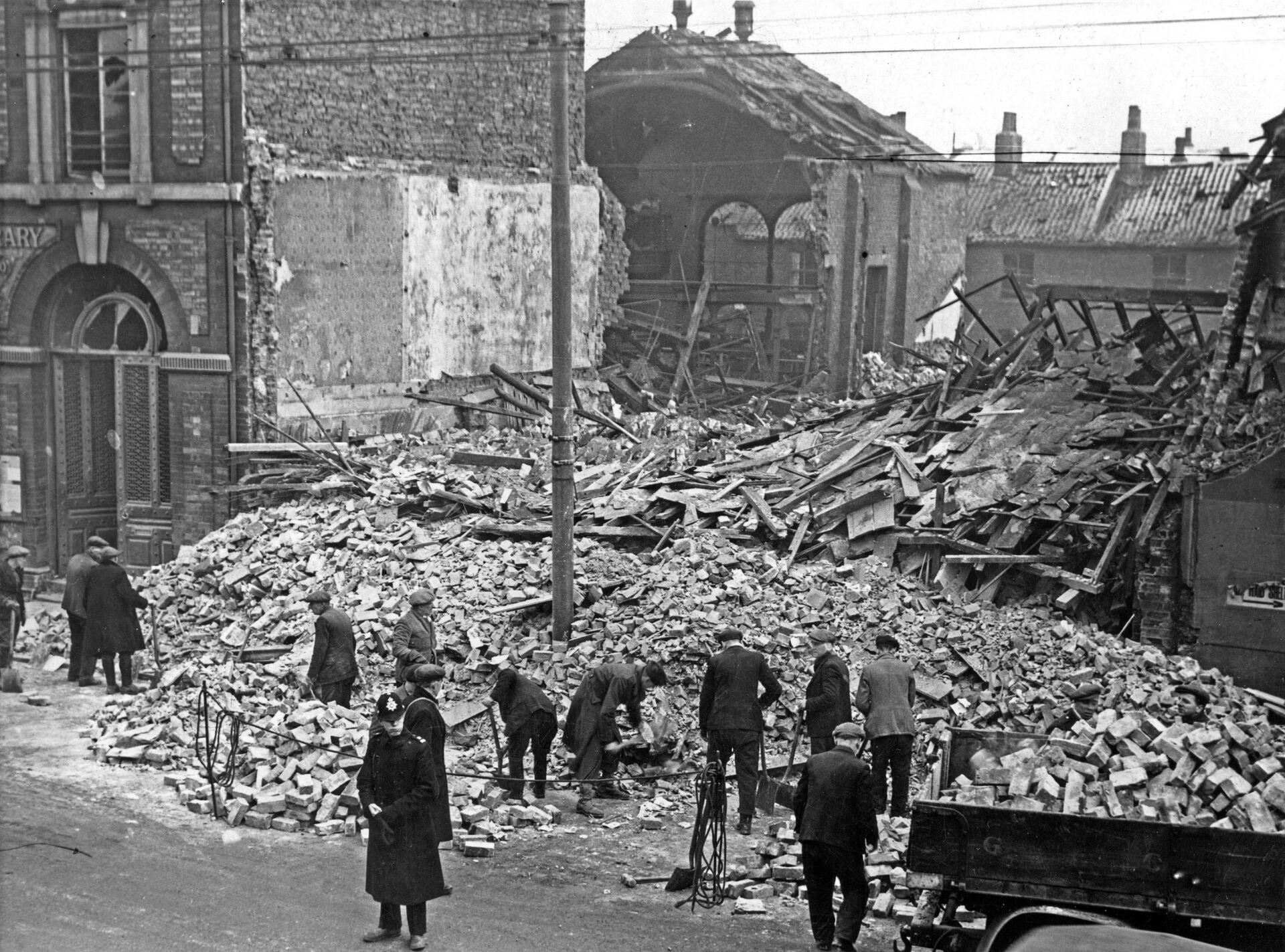Debris from bombed buildings, people search rubble. Brick, concrete, and damaged structures are visible.