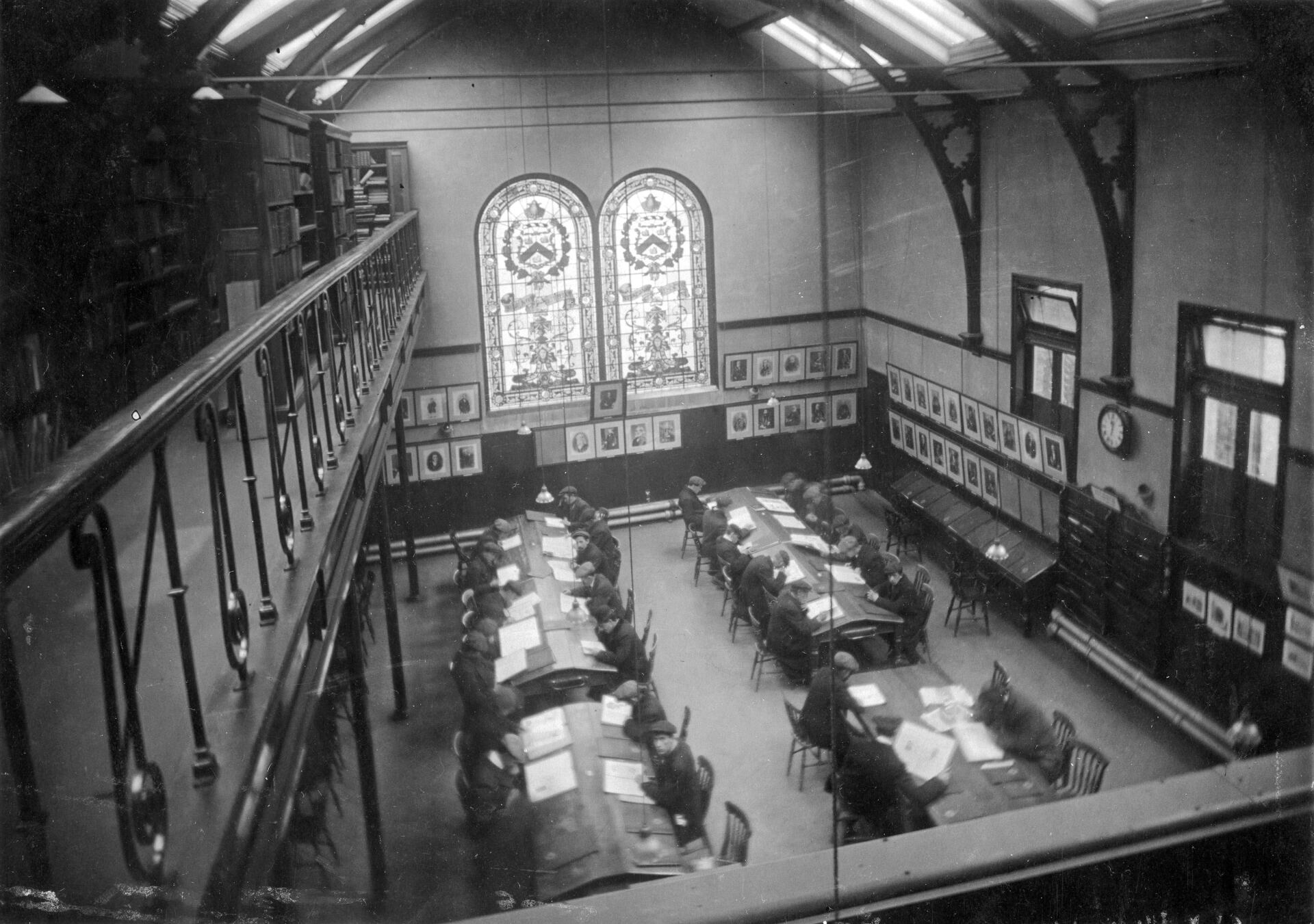 High-angle view of a library with tables and chairs for reading. Two large stained-glass windows are centered.