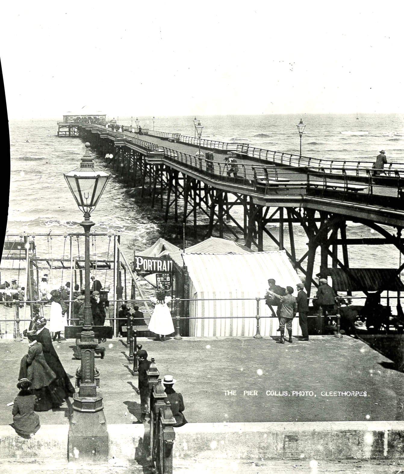 Old pier extending into the sea; people on the pier and along the shore in front of some structures.