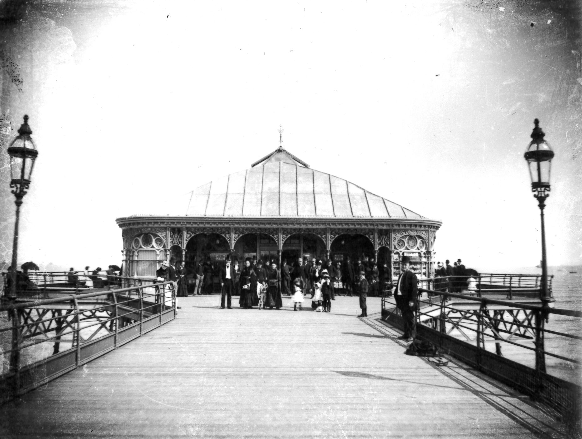 Pier with people gathered at a pavilion. Ornate railings and lampposts frame the wooden walkway.
