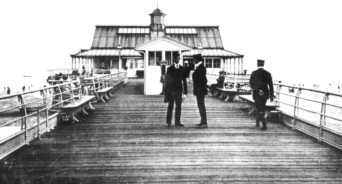 Two men in suits converse on a wooden pier with benches, a building in the background.