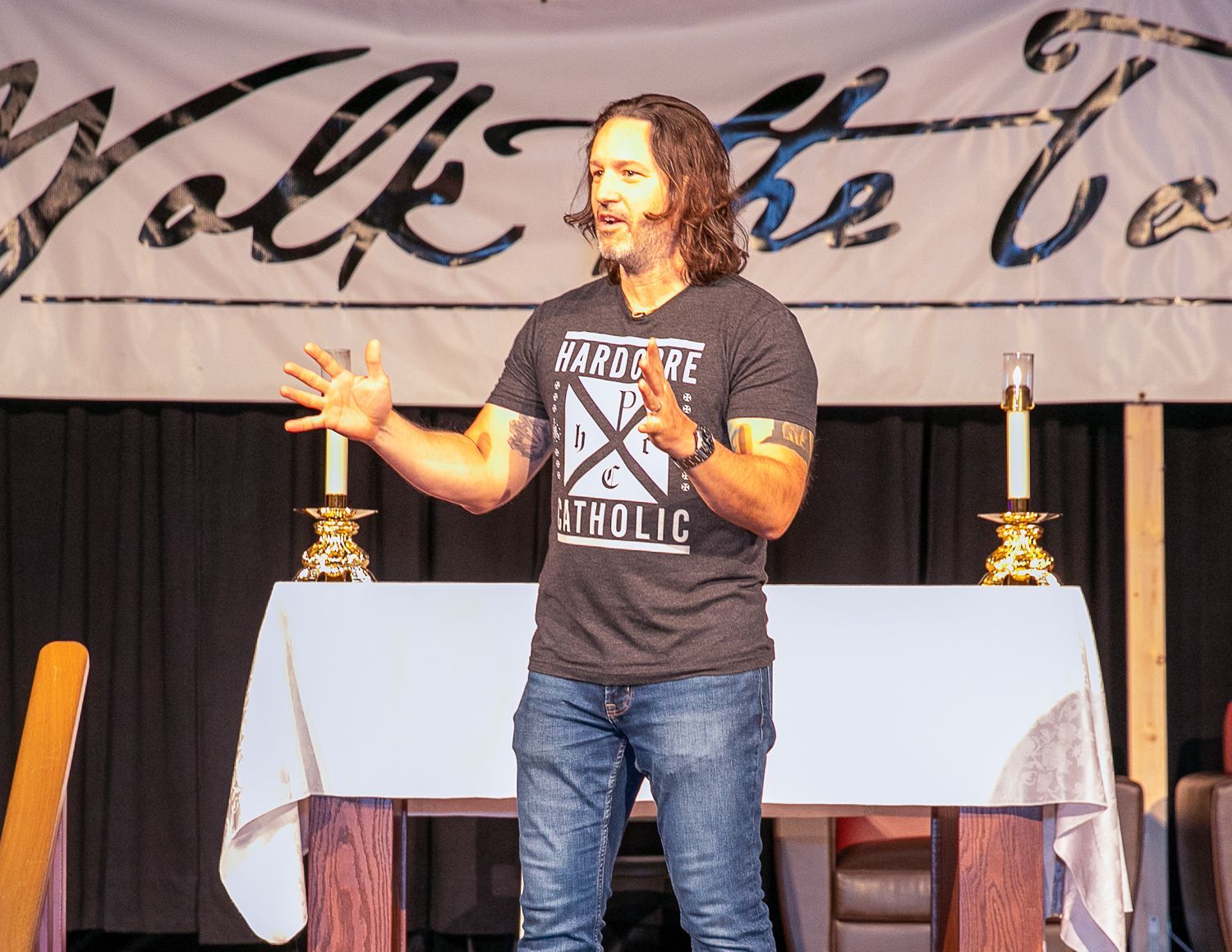 Keith Nester speaking on stage beside a table with candles, in front of a banner backdrop