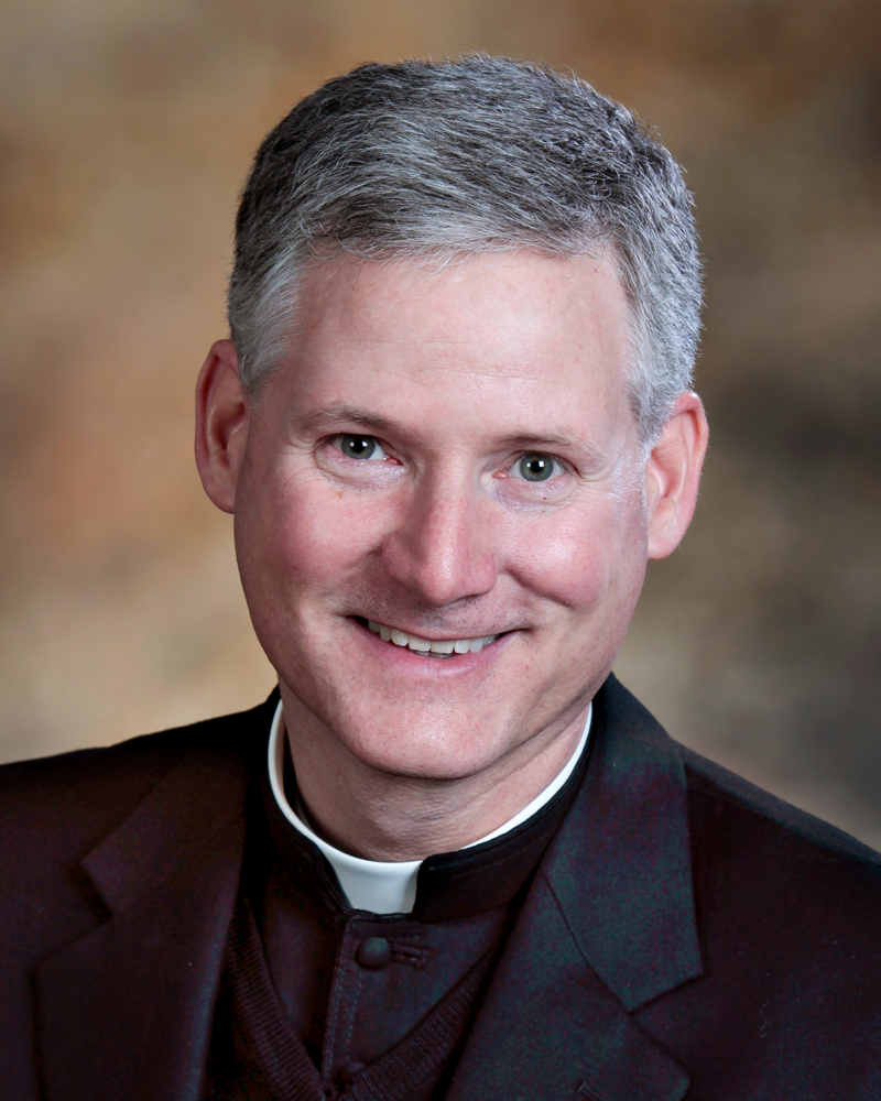 Portrait of Fr. Rocky smiling in a dark suit and clerical collar against a warm studio background
