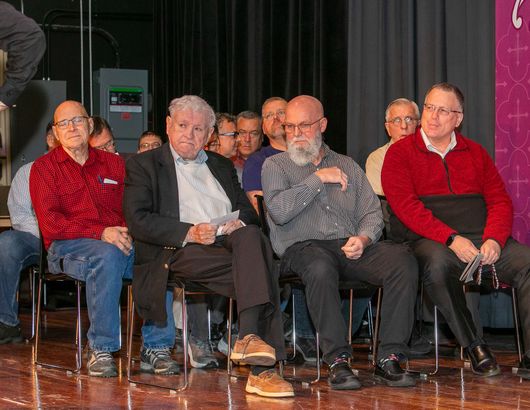 Six men seated on stage in chairs, wearing casual to semi-formal clothes, listening during an event.