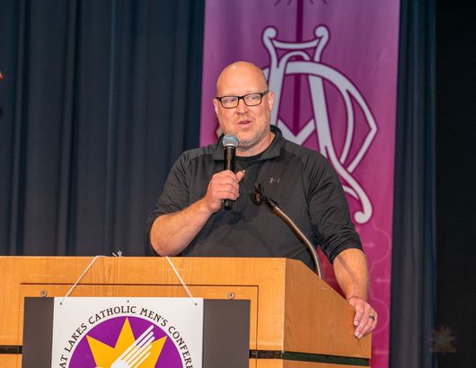 Mitch Striedl speaking at a podium on a stage with a purple backdrop and black curtains