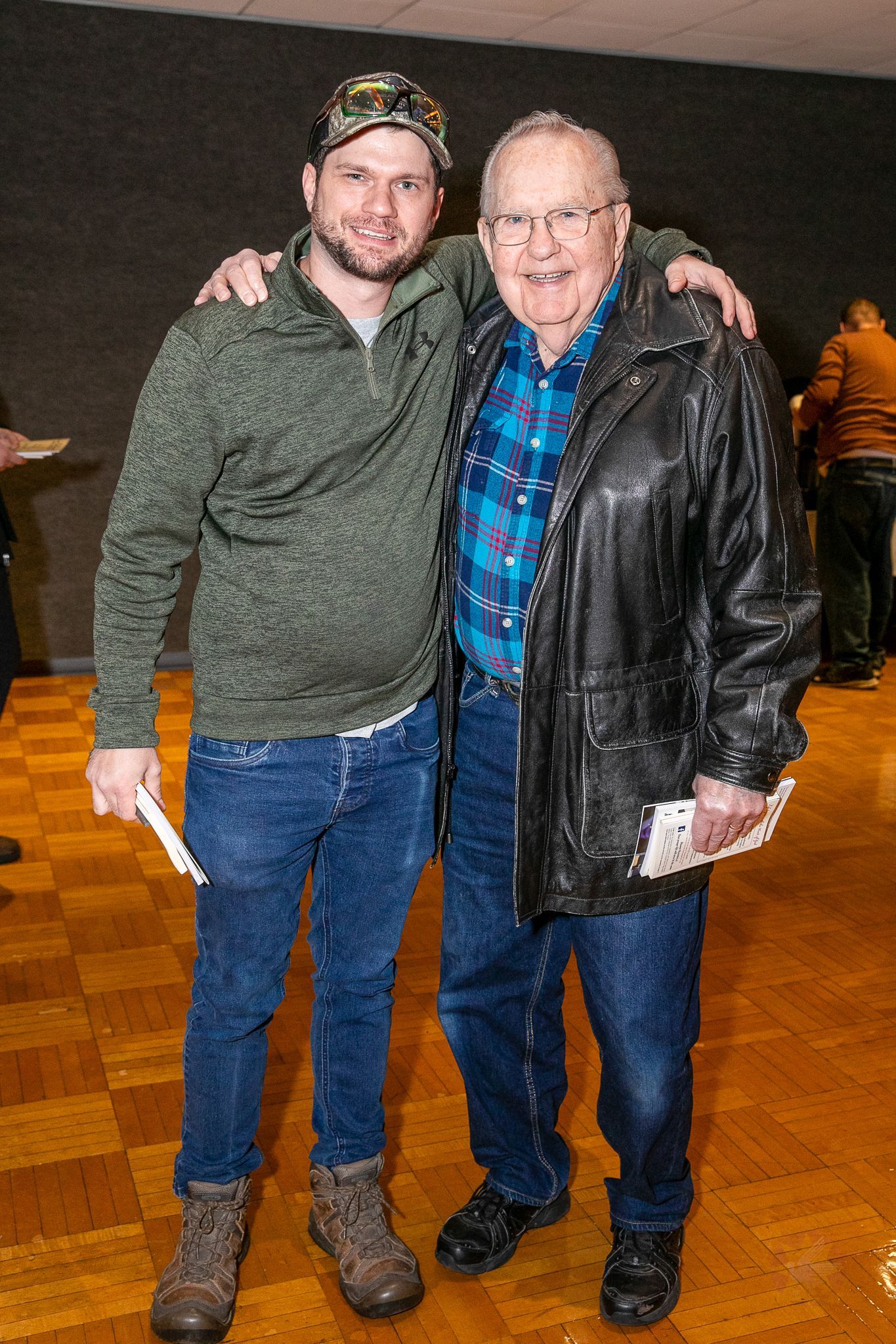 Two men standing together indoors, smiling, one in a gray sweater and cap, the other in a black jacket and blue shirt