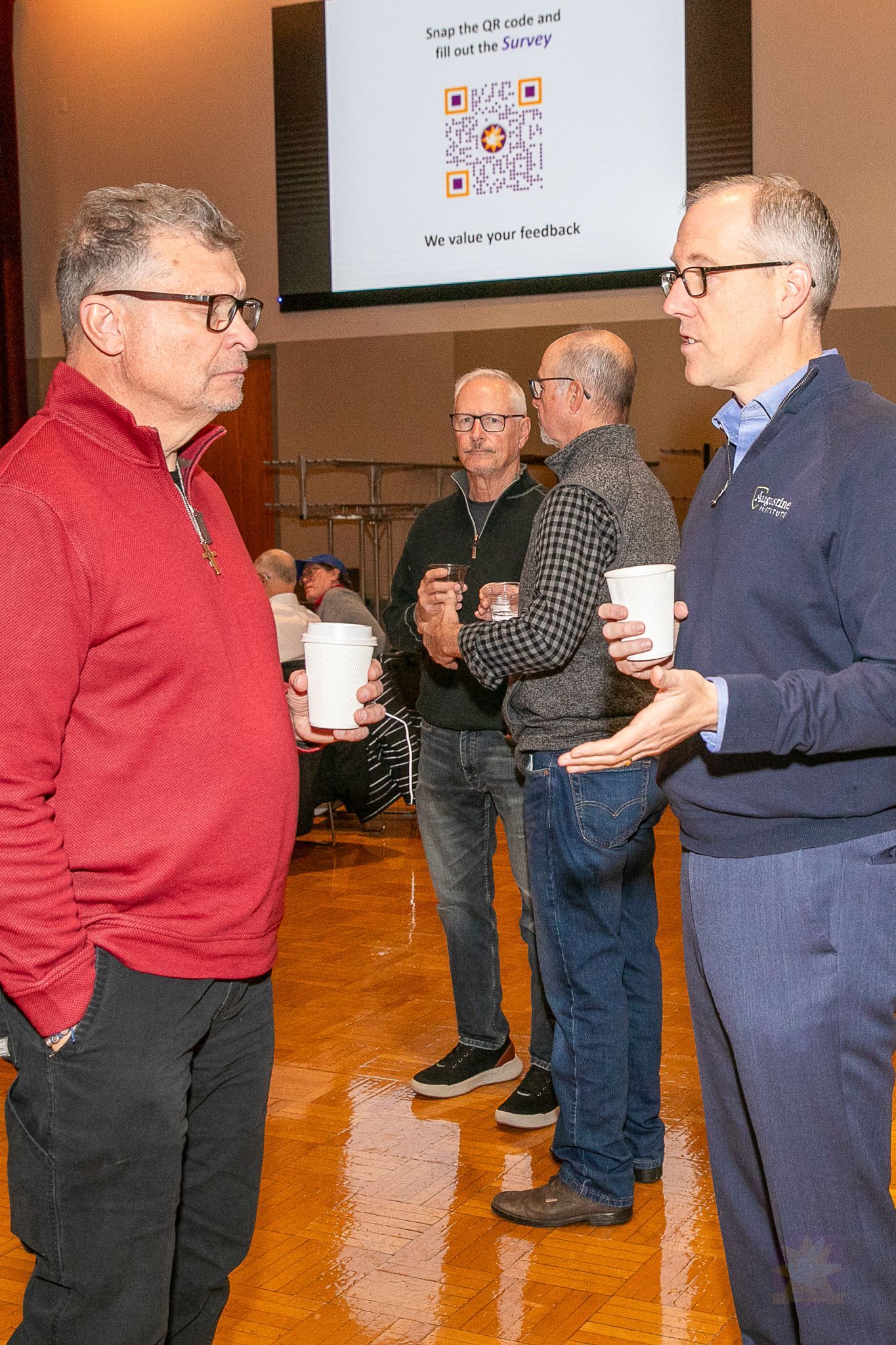 Three men talking at an indoor event, holding cups, with a presentation screen in the background.