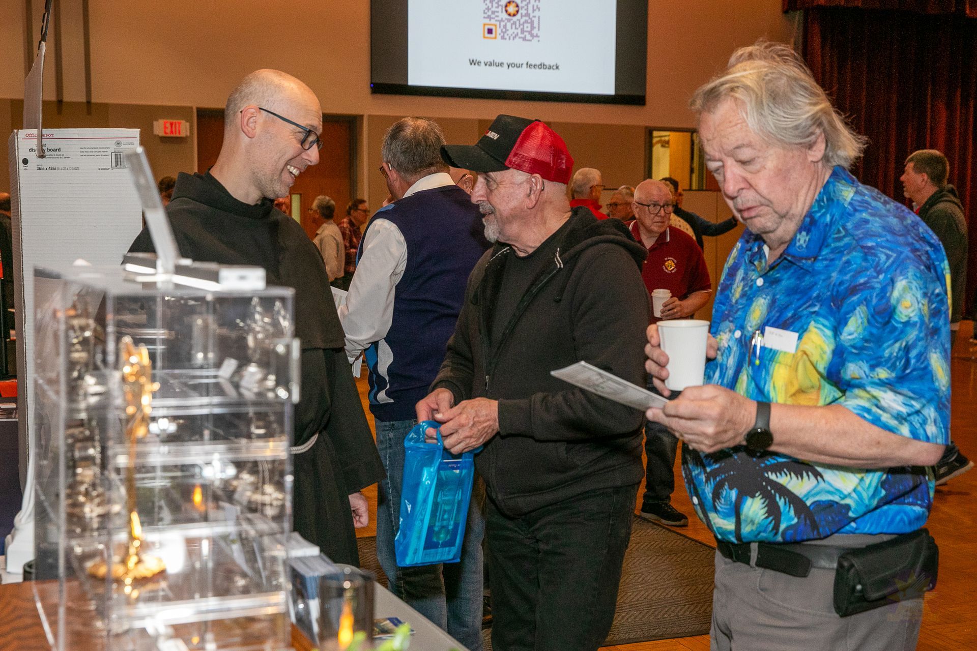 People browsing at a Catholic men's conference, with a man in a blue Hawaiian shirt reading a paper.