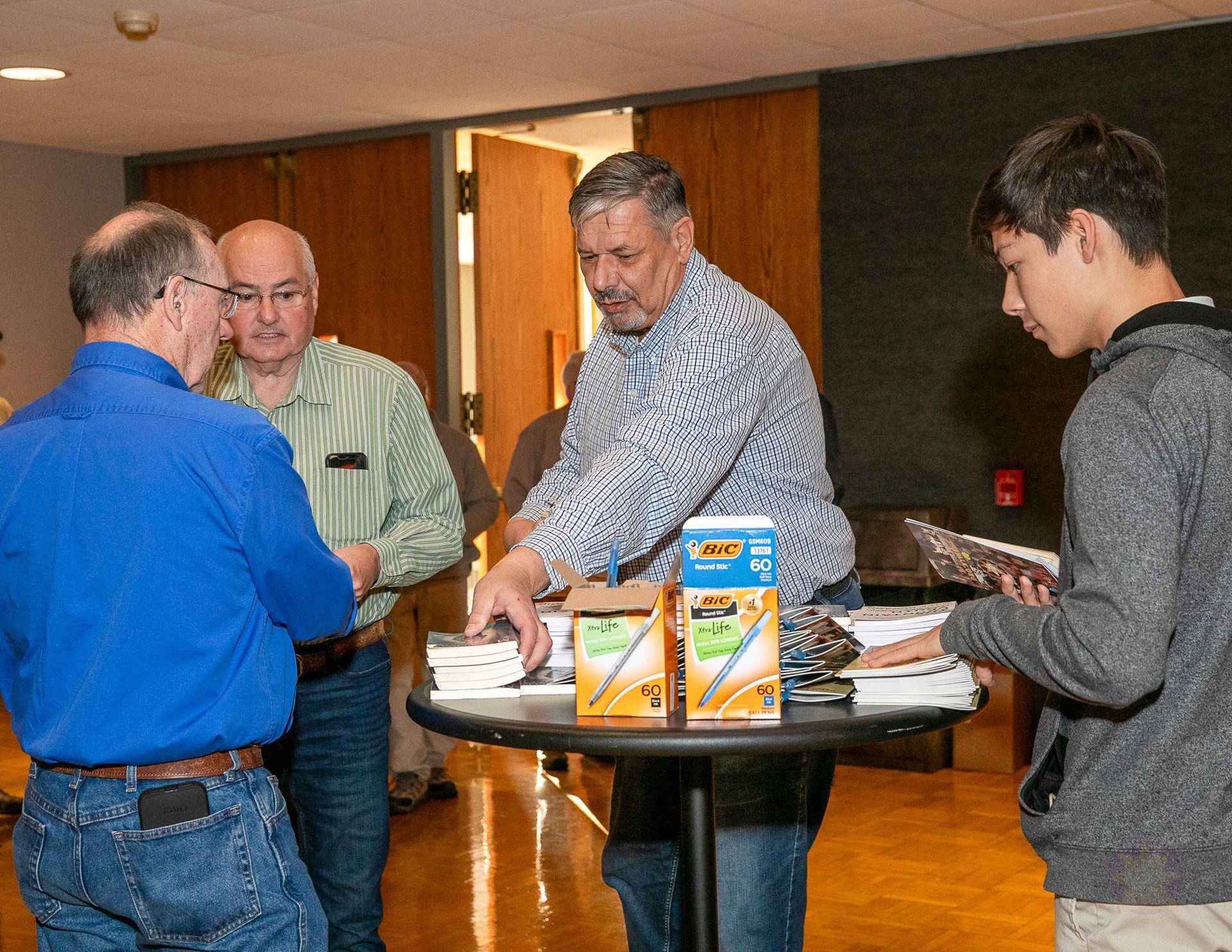 Four people converse around a round table with brochures and papers in a warm indoor lobby.