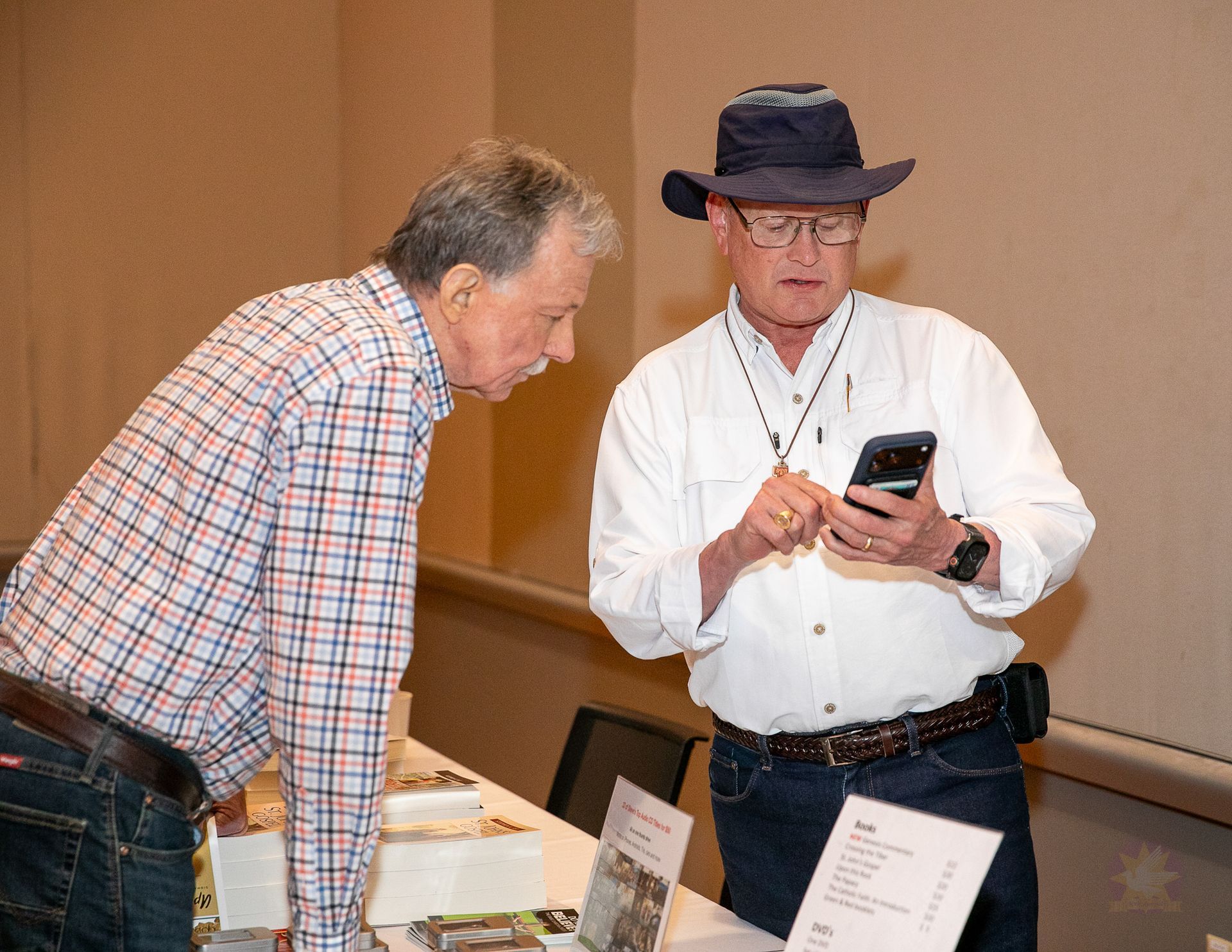 Two men at a table; one in a hat checks a phone while the other leans over, with display items nearby.