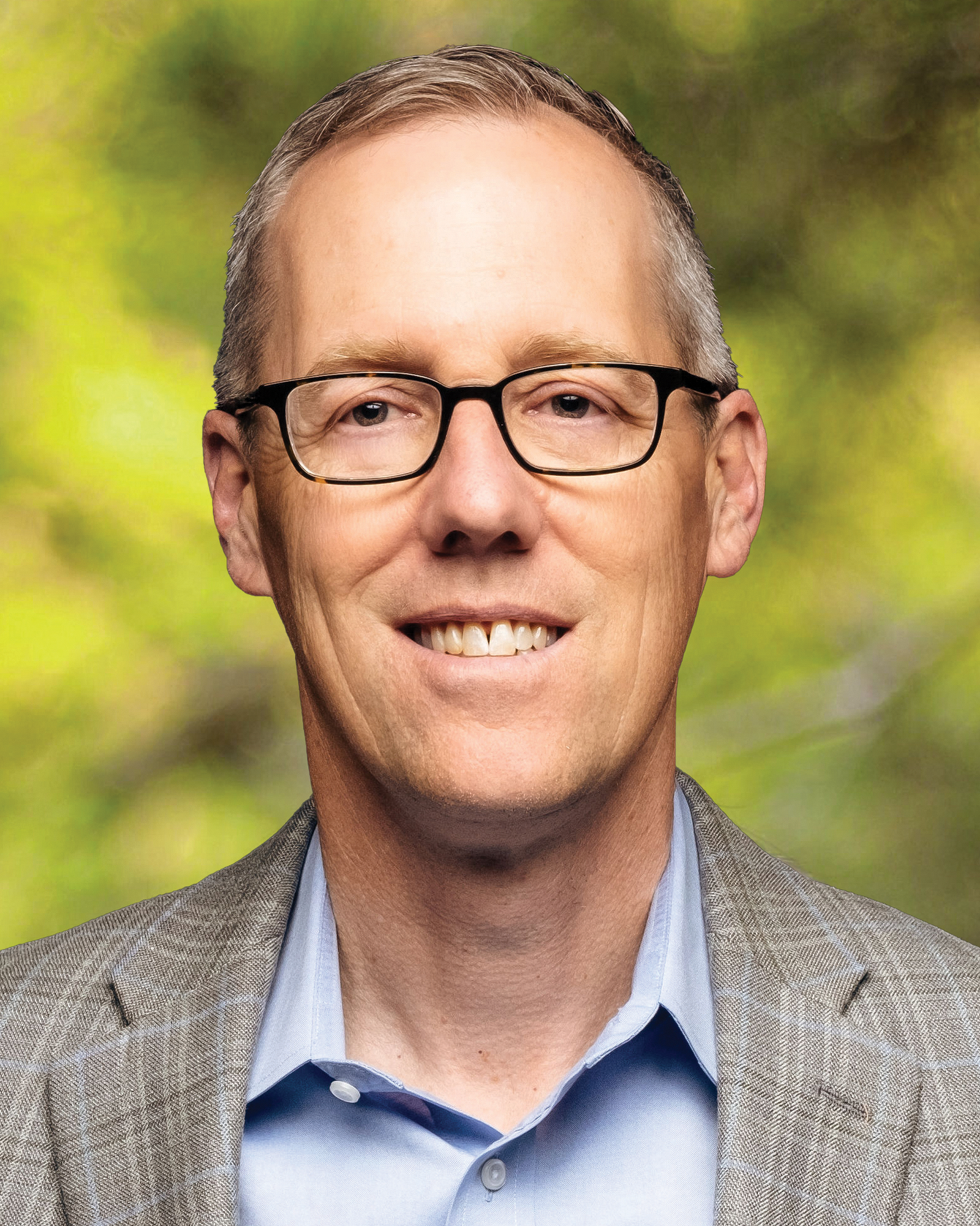 Portrait of Dr. Tim Gray smiling, wearing glasses, a blue shirt, and a gray blazer against a blurred green background