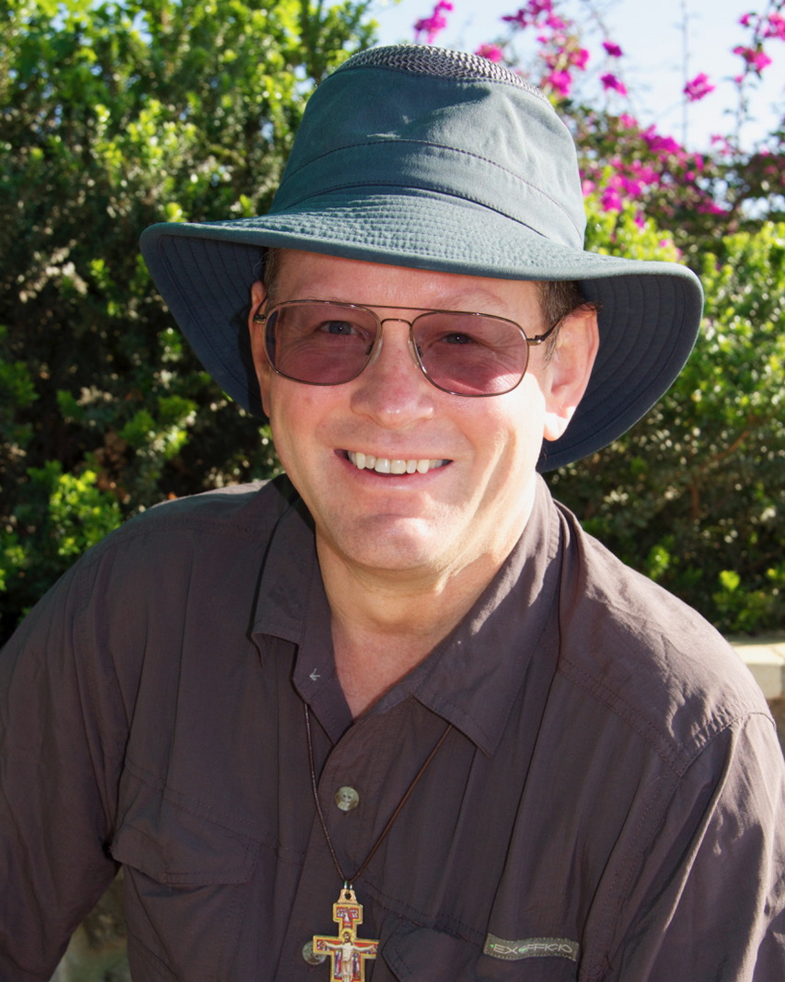 Portrait of Steve Ray smiling in a dark hat and sunglasses outdoors with green bushes and pink flowers.