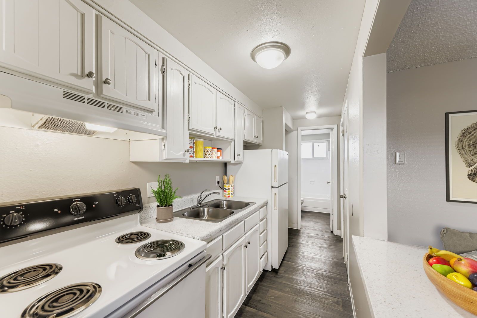 Bright galley kitchen in an apartment with white cabinets, stove, double sink, and refrigerator.
