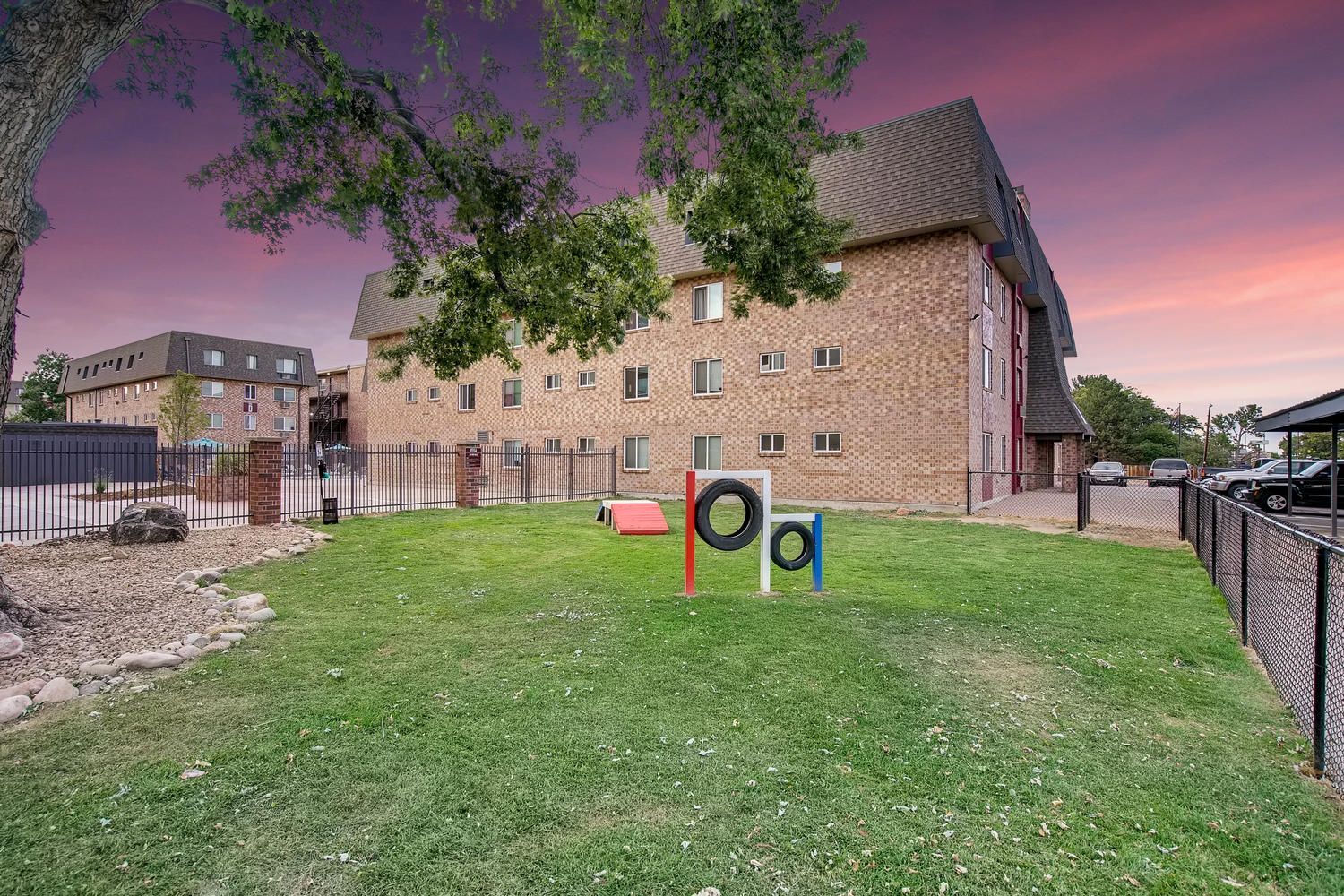 Exterior view of a brick apartment building with a fenced green lawn and small tire play equipment.