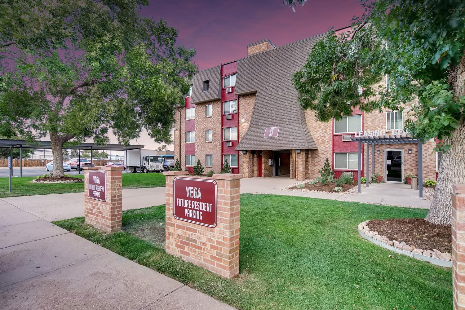 Exterior view of Vega apartment complex with leasing center and signs, trees, and parking.