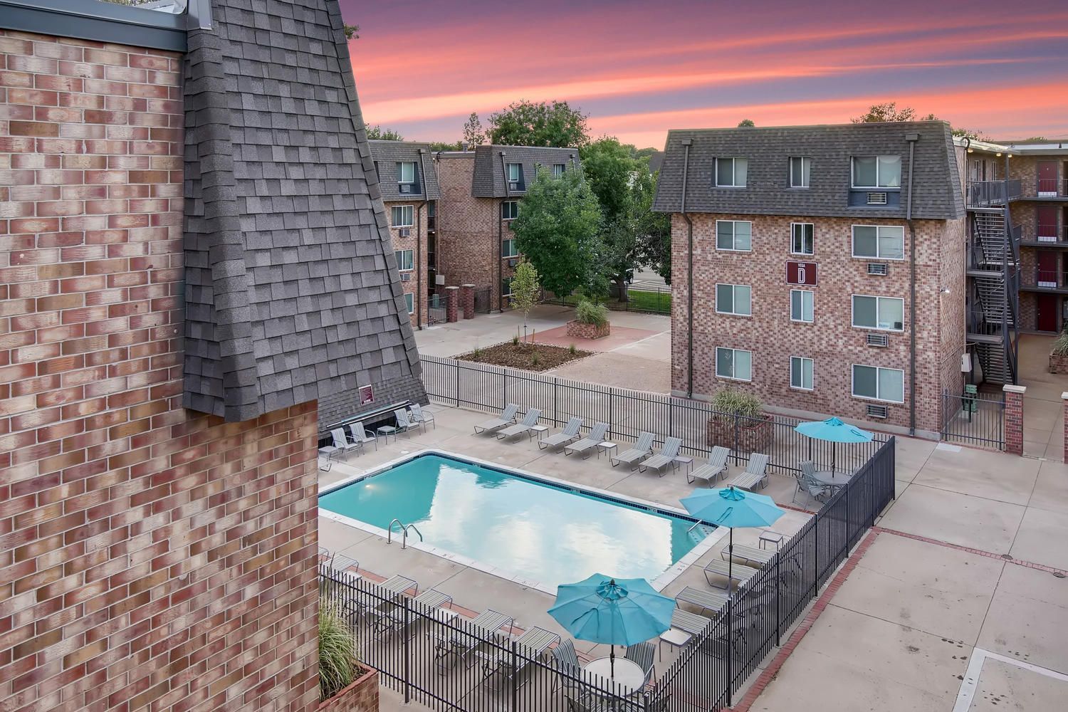 Apartment complex with a pool, lounge chairs, and buildings. Red brick exterior with a sunset sky.