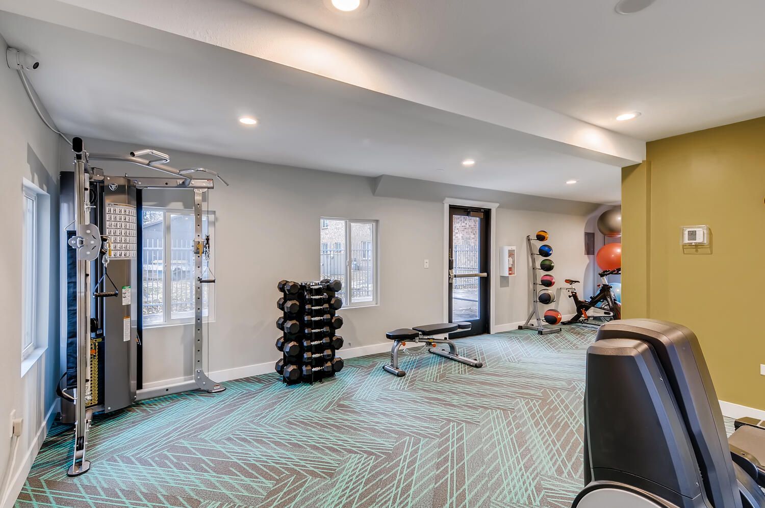 Indoor community gym with free weights, bench, dumbbells, and cardio machines.