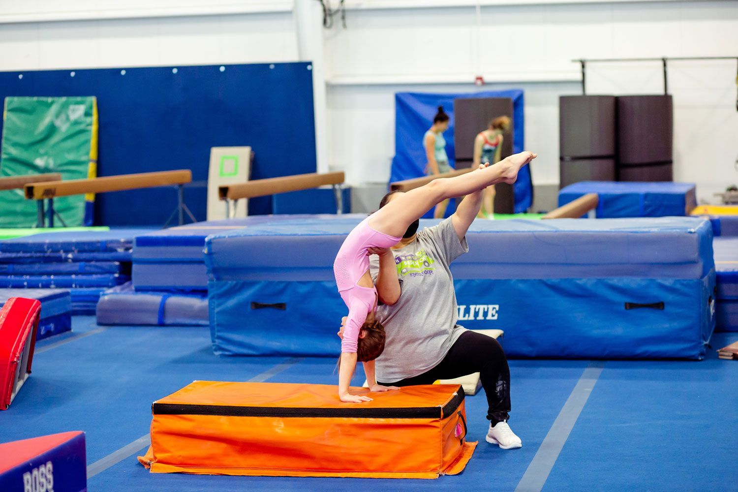 A woman is helping a young girl do a handstand on a mat in a gym.