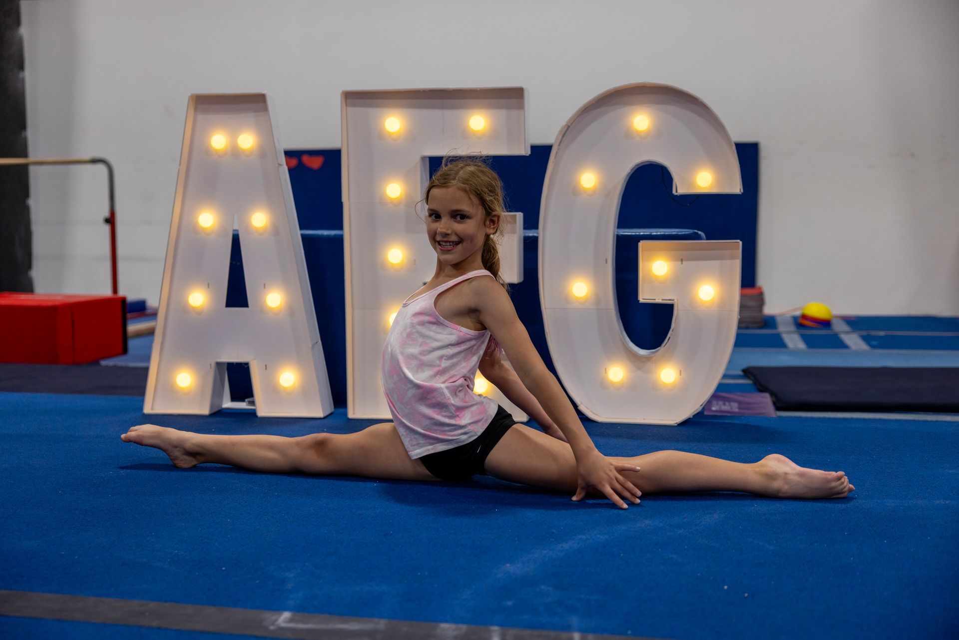 A young girl is doing a split on the floor in front of a sign that says afg.