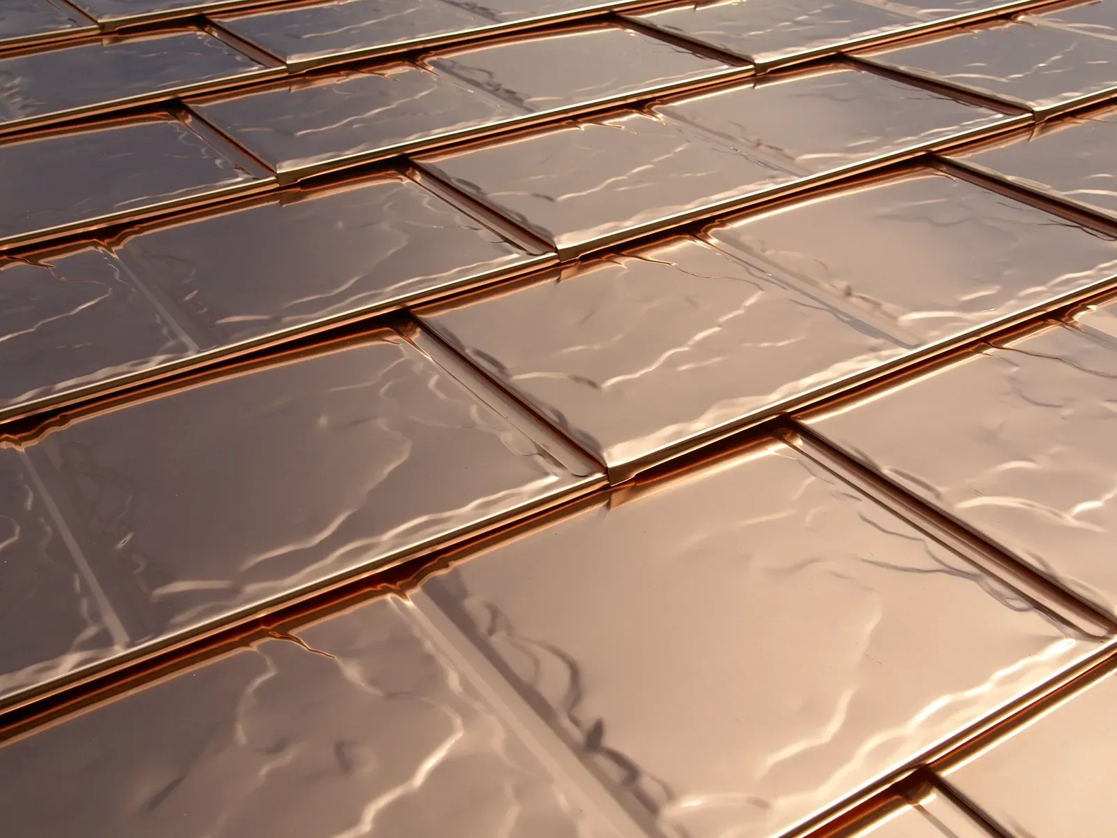 A close-up, angled view of rectangular, copper-colored metallic roofing tiles with a rippled, reflective surface texture.