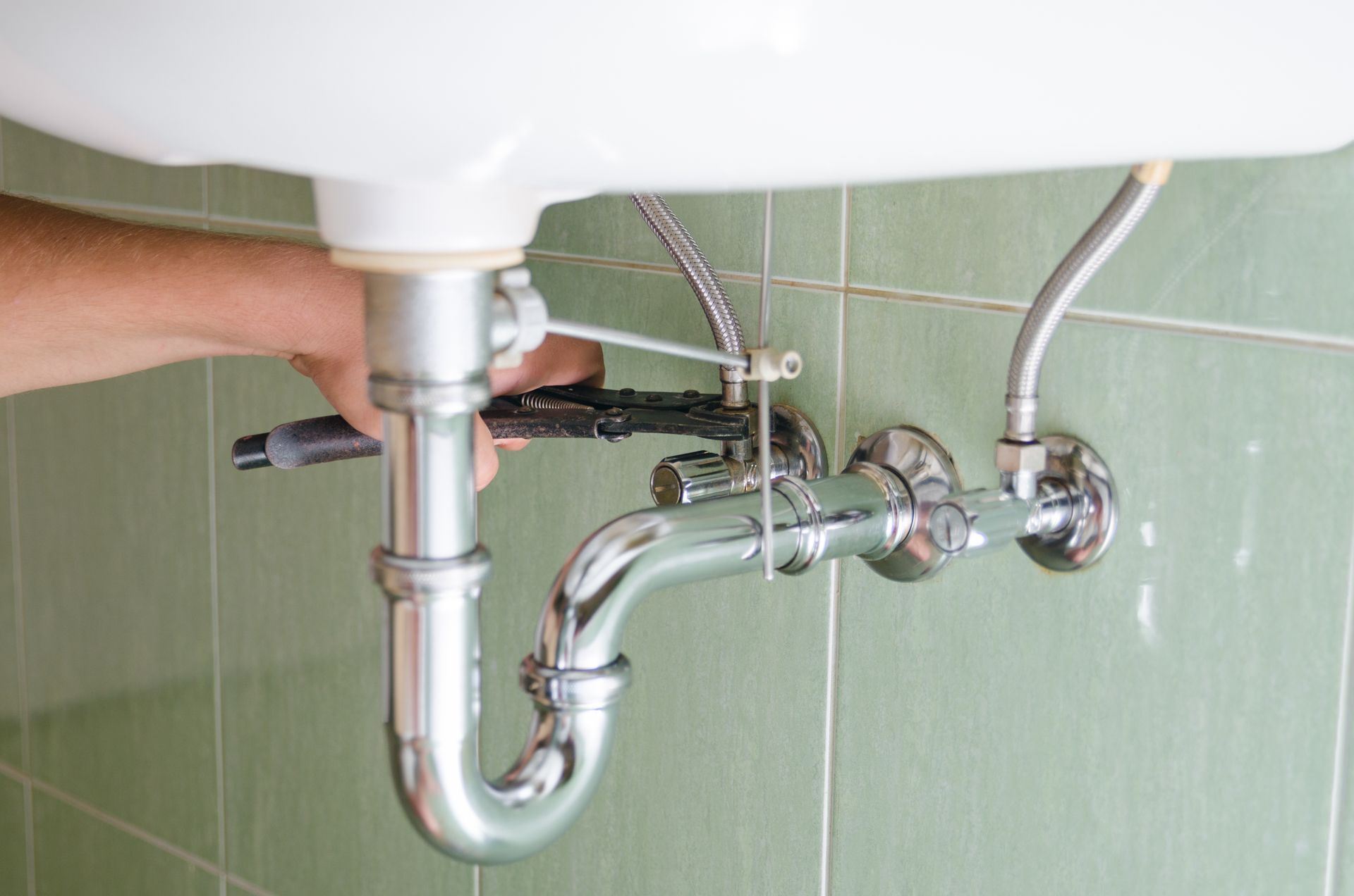 A person using a wrench to tighten the plumbing under a white sink against a green-tiled wall.