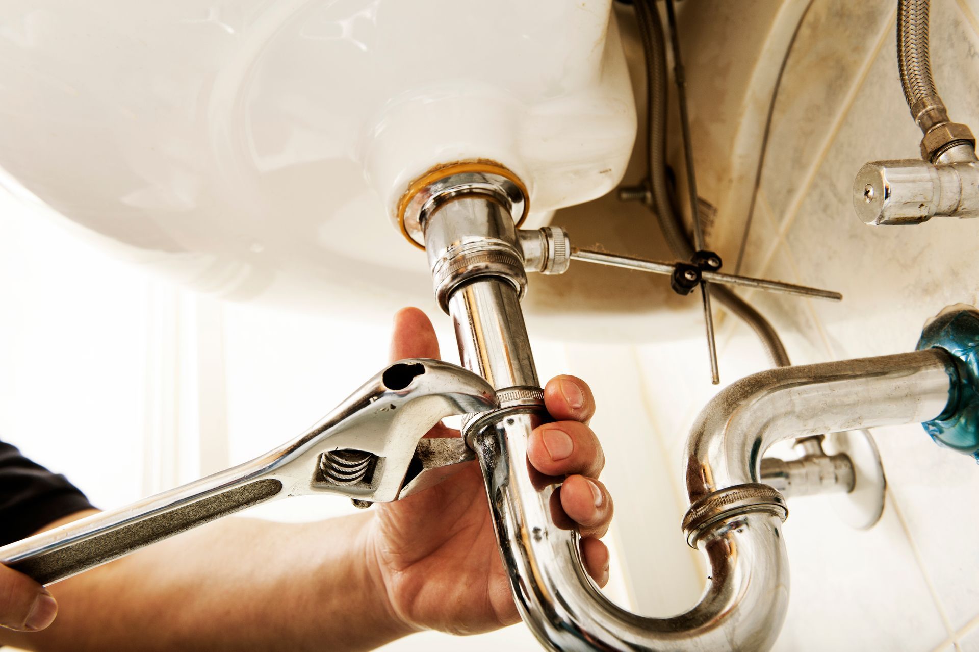 A plumber using a wrench to tighten a pipe under a white sink.
