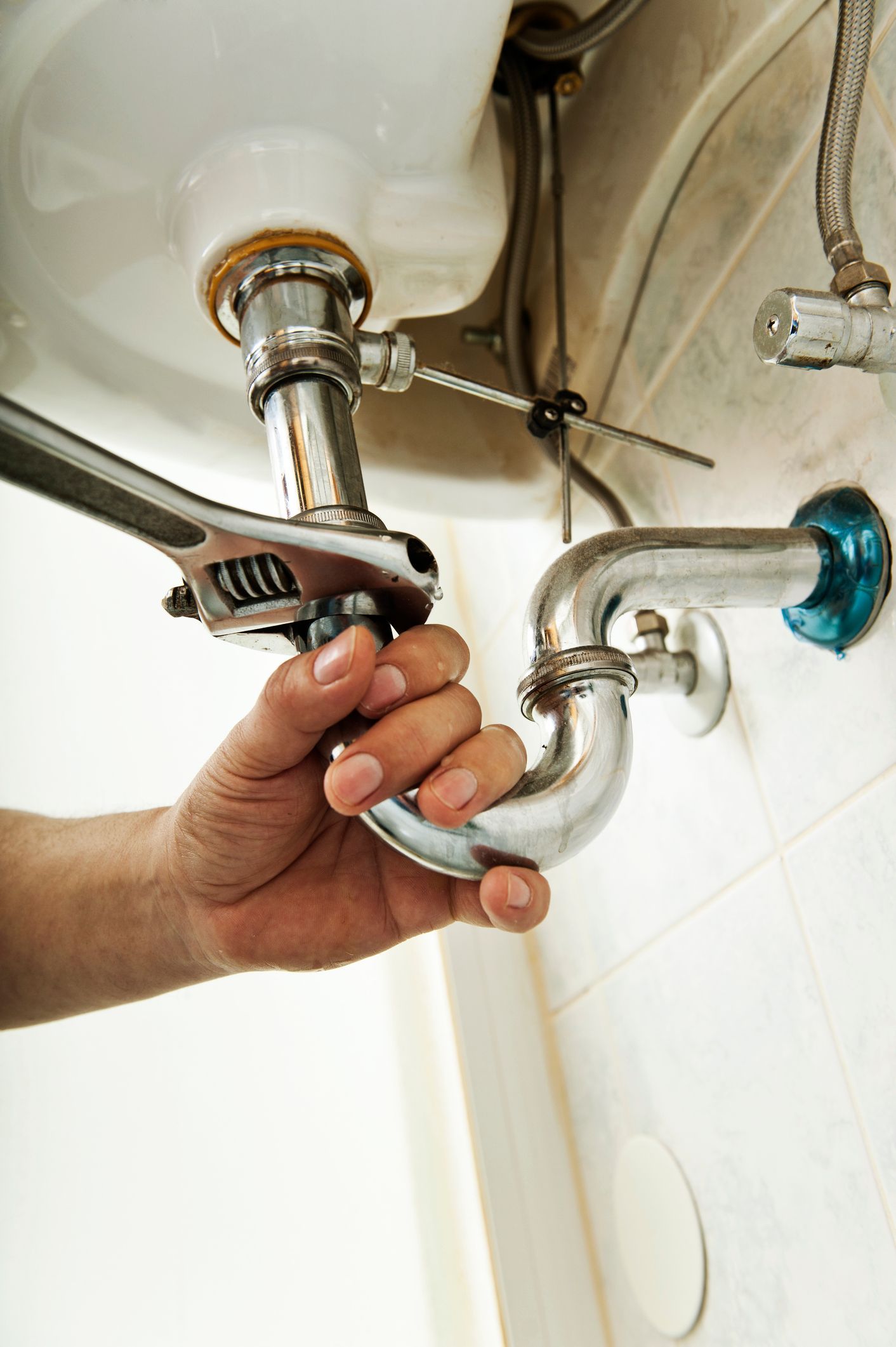 A person's hand uses a wrench to tighten a curved pipe under a white sink in a bathroom.