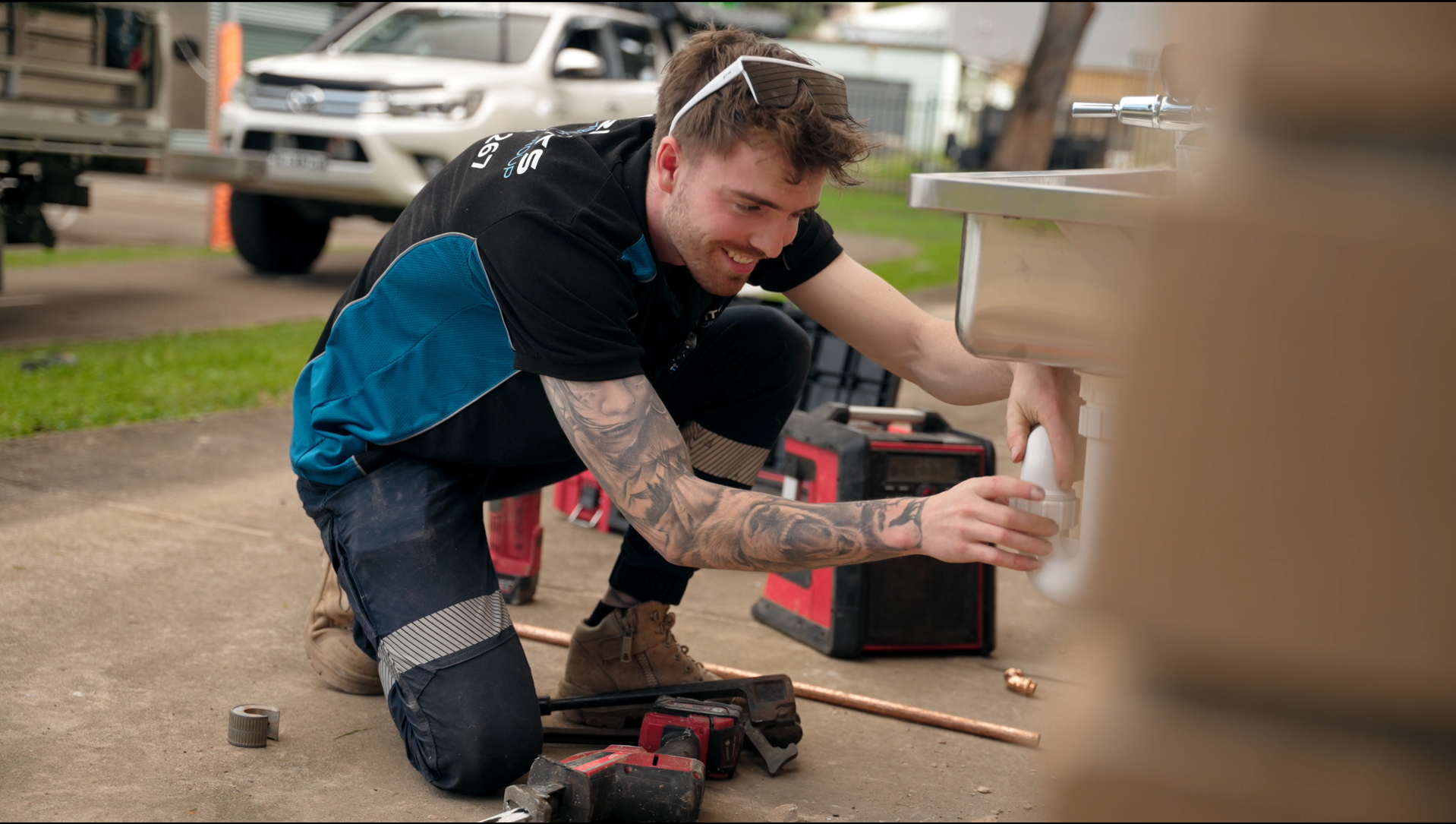 Plumber working under a kitchen sink with a red adjustable wrench. Black pipes and chrome sink are visible.