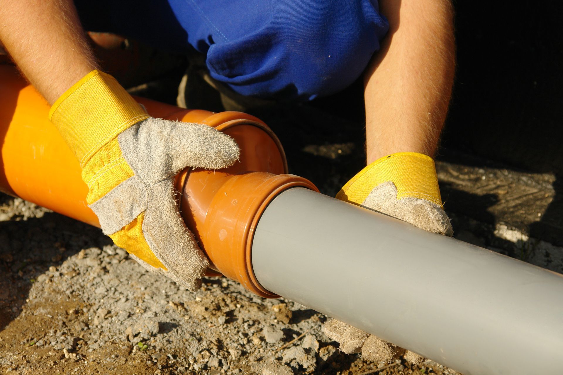 Hands wearing yellow gloves connect a brown pipe fitting to a gray pipe outdoors.