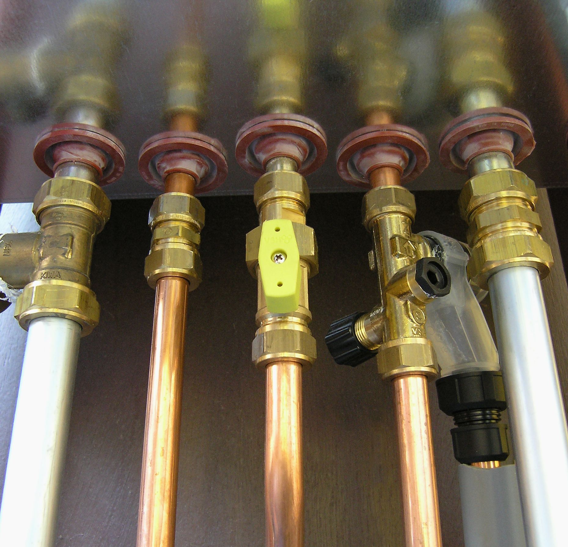 Close-up of copper pipes with brass fittings and valves, likely part of a plumbing or heating system. Some pipes are connected to white plastic tubing.