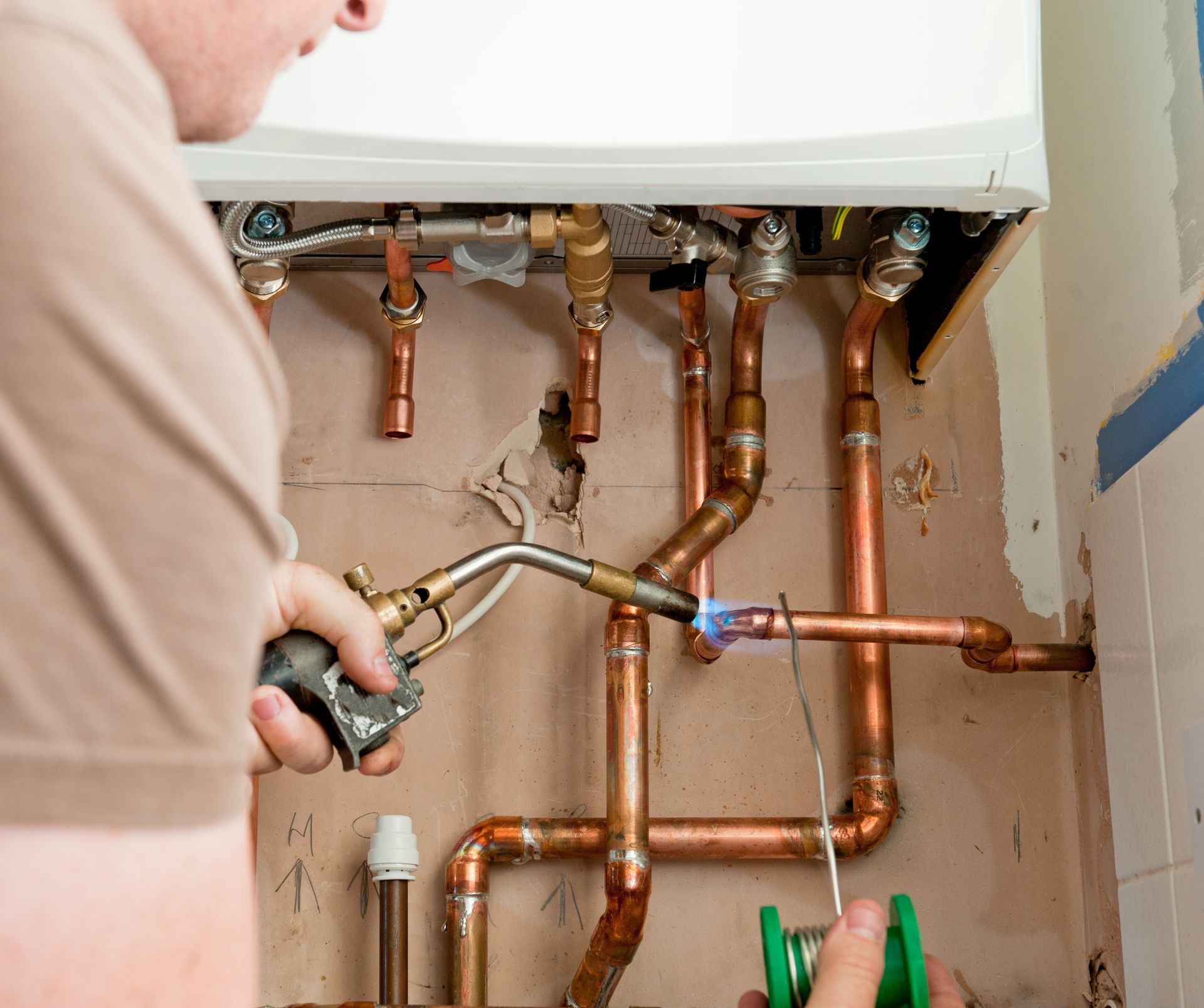 A plumber using a torch to solder copper pipes connected to a white appliance on a wall.