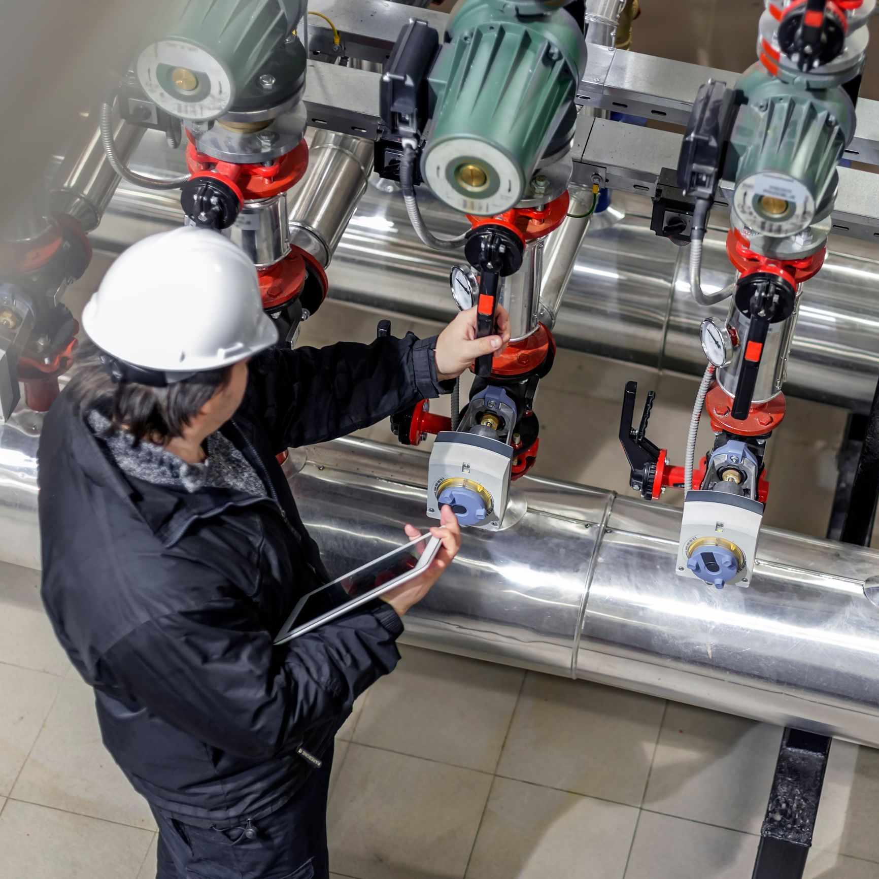 Engineer in hard hat examining industrial equipment with a tablet, near pipes and machinery.
