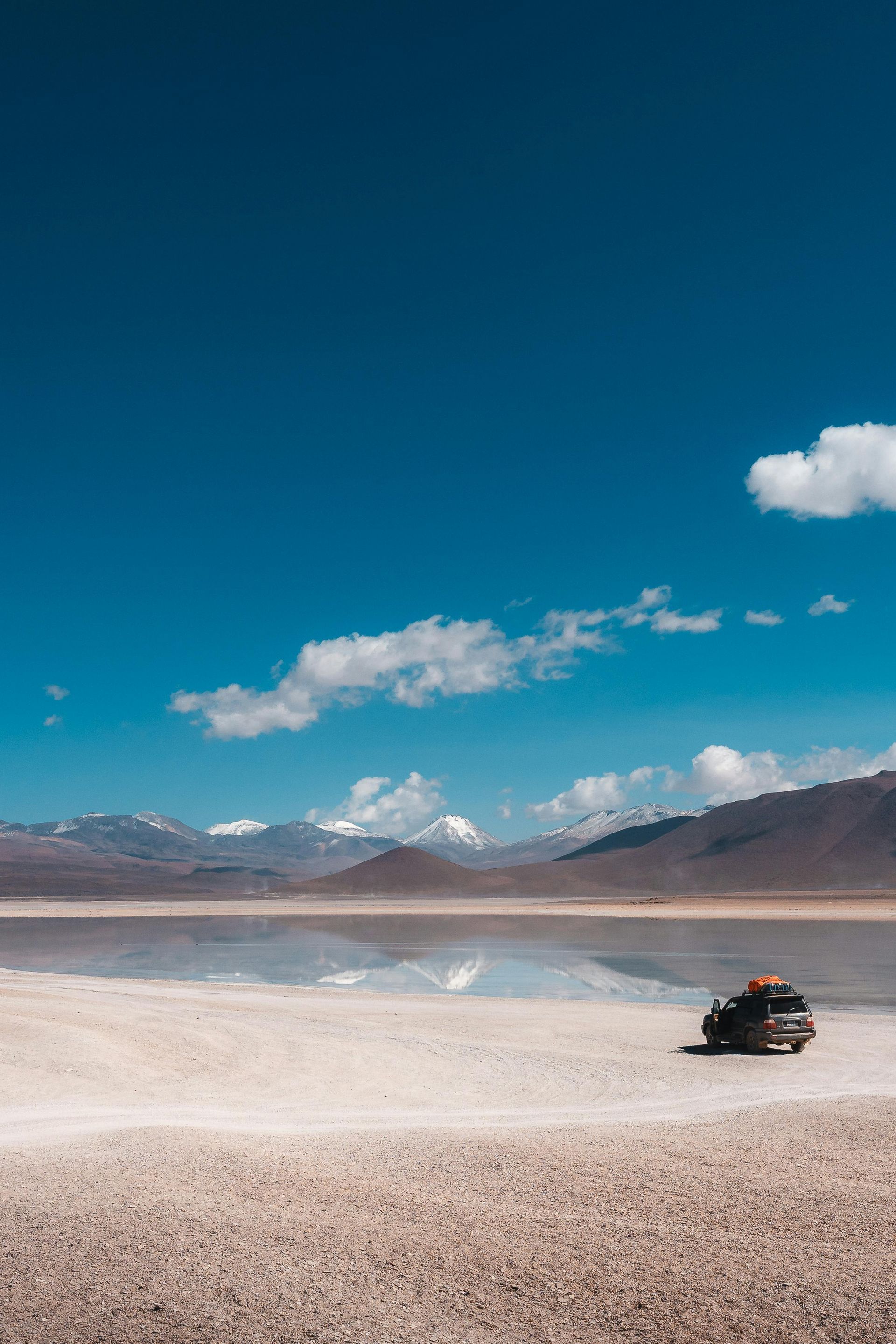 A car is parked on the shore of a lake with mountains in the background.