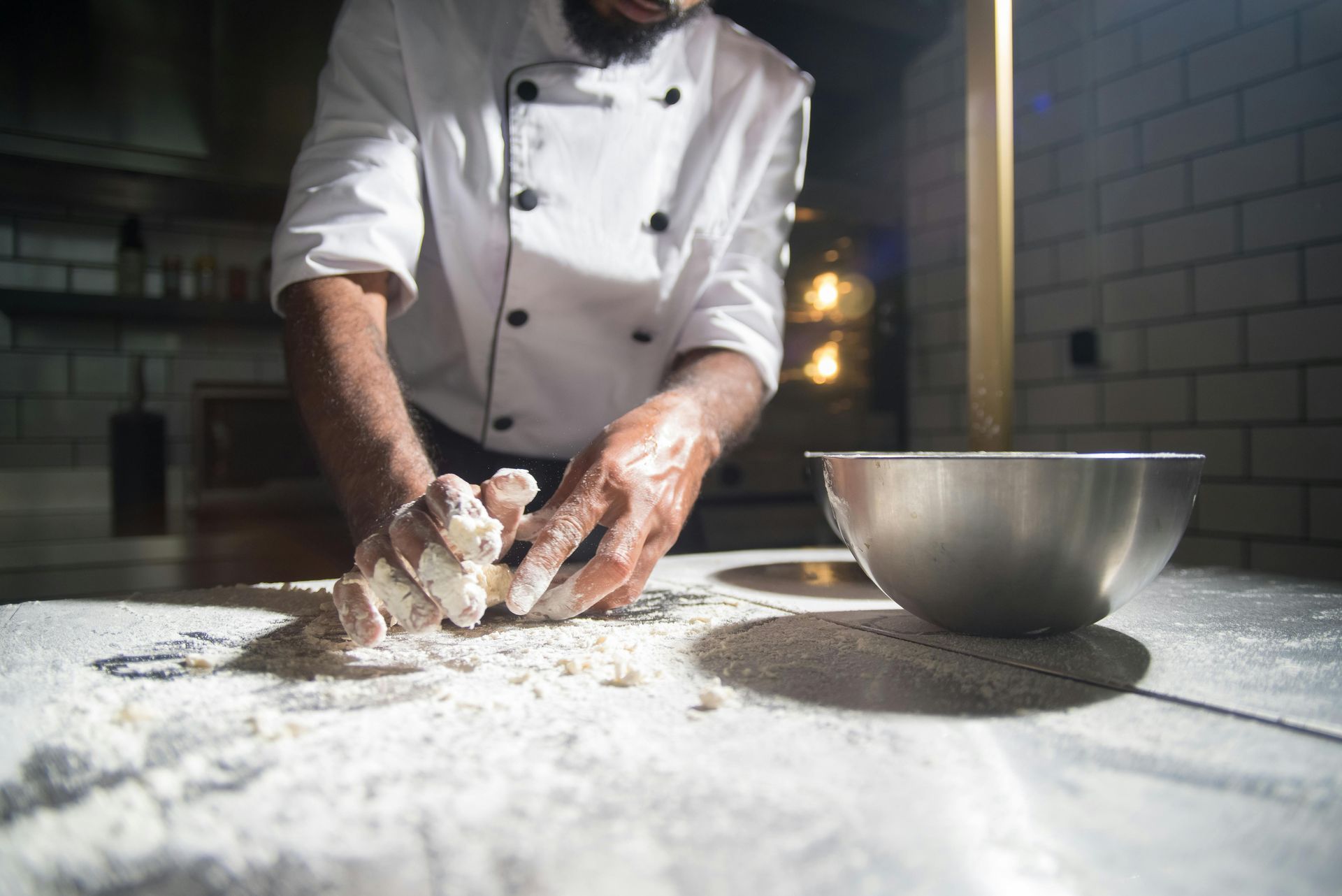 Chef kneading dough on a floured countertop near a metal bowl, wearing a white uniform in a kitchen.