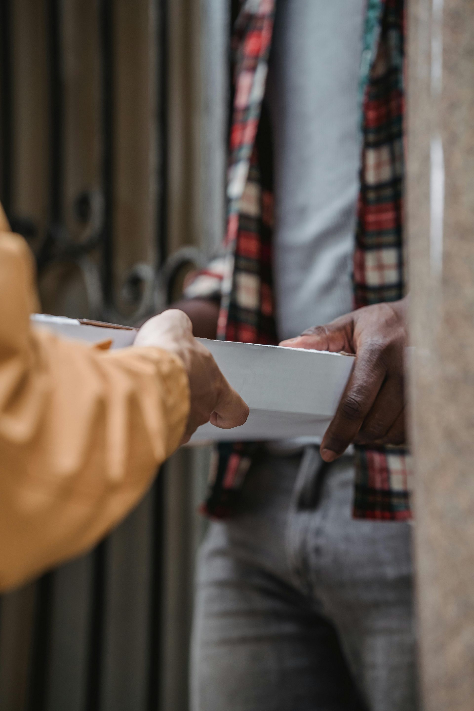 Two people exchanging a white takeout container. One person wears a tan jacket, the other, a plaid shirt and jeans.