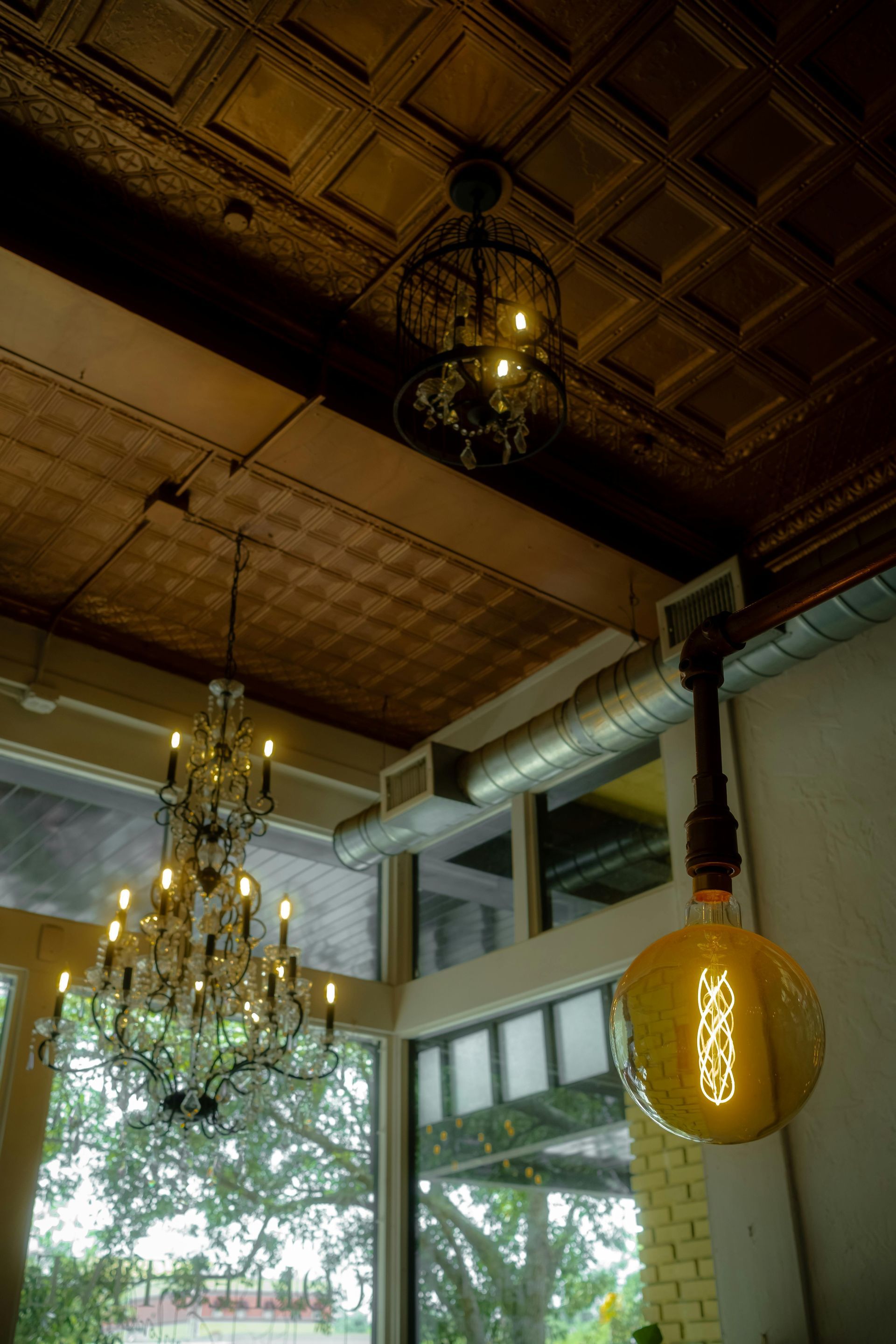 Ornate chandeliers hang in a room with a patterned ceiling, industrial light fixture, and large windows.
