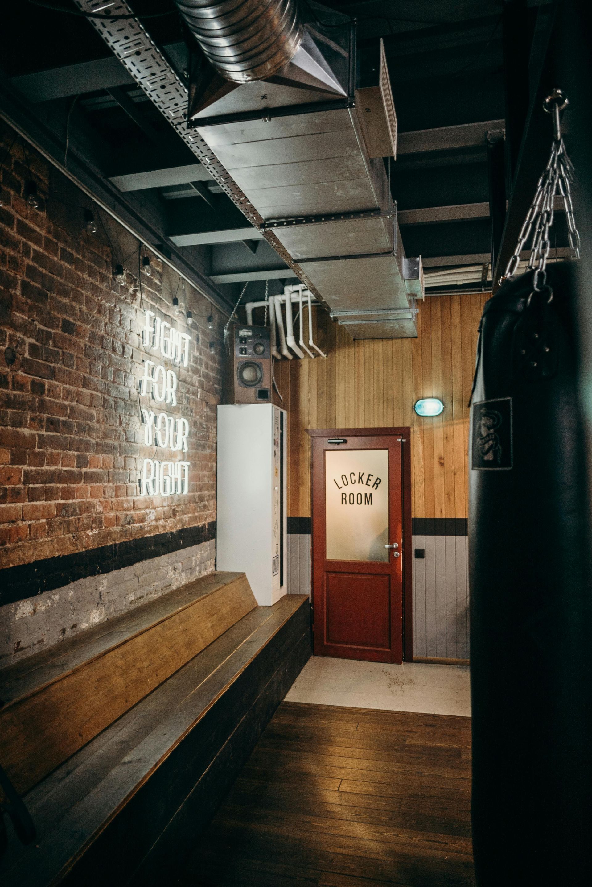 Narrow gym hallway with exposed brick, wooden bench, punching bag, and doorway. Neon sign on brick wall.