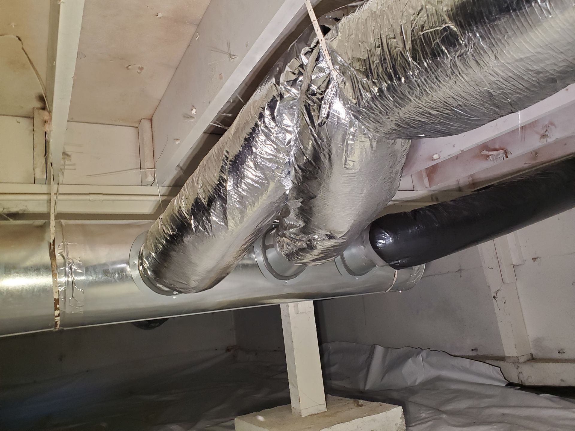 Ductwork in a crawl space, with silver-wrapped insulation and a black pipe, under a wooden structure.