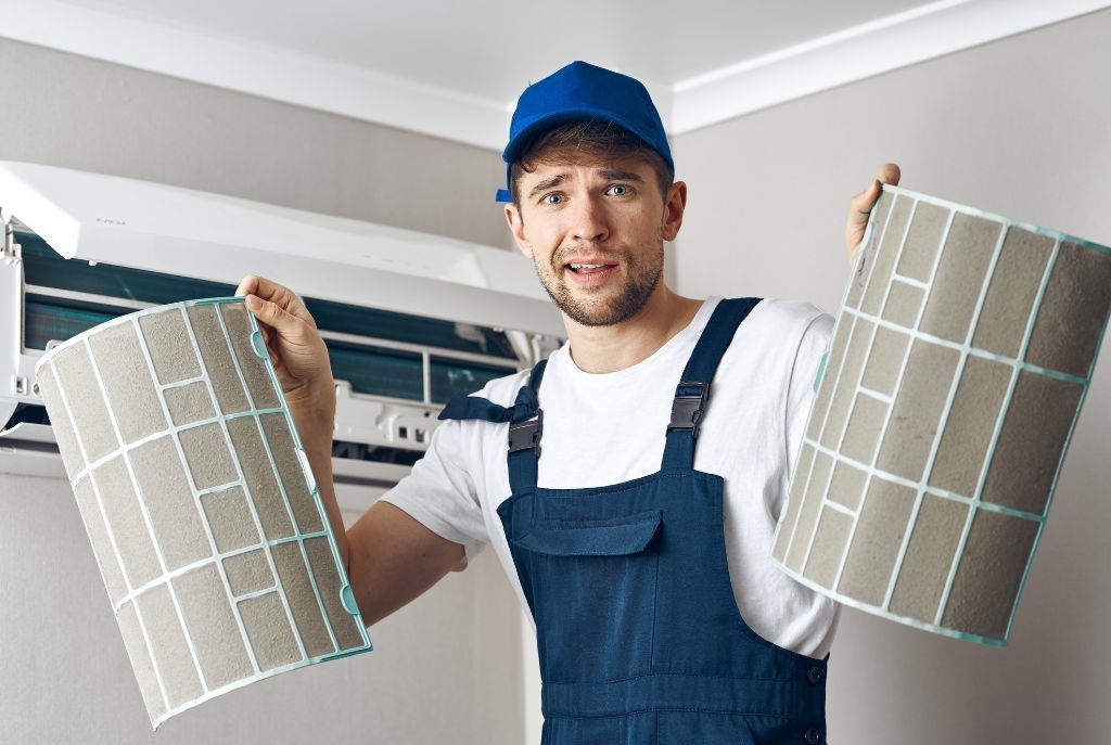 A man is holding two dirty filters in front of an air conditioner.