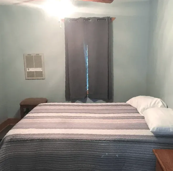 A bedroom with light blue walls, featuring a bed with a gray striped comforter and white pillows, next to a small table.