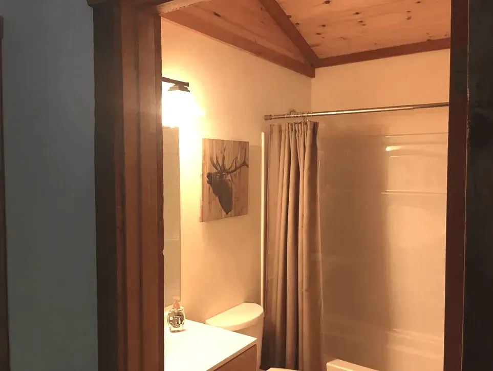 A bathroom viewed through a doorway, featuring a vanity with a mirror, a toilet, a shower with a tan curtain, and wood ceiling.